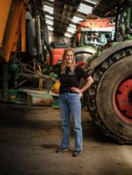Dame die in de loods staat met een veiligheidshelm onder haar arm. Op de achtergrond tractoren.